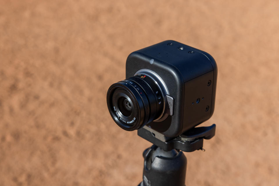 Lifestyle marketing photo of the Logitech Mevo Core live streaming camera.  Seen slightly from above, facing its front left side.  He is mounted on a tripod with a field of earth (blurred) visible behind him.