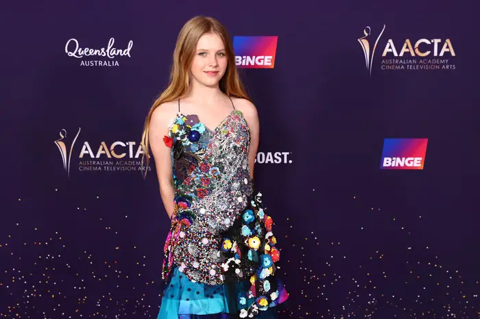 Alyla Browne wears a bright, floral, blue dress and poses in front of a sign bearing the name and logo of the Australia Academy Cinema Television Arts.
