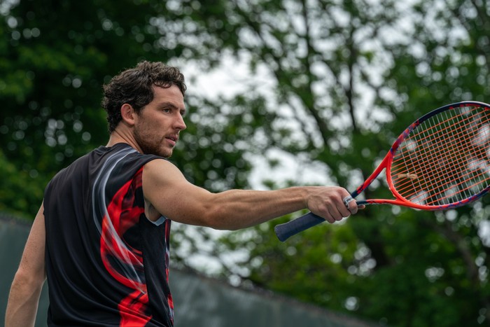 Josh O'Connor plays Patrick wearing a red and black vest and holding a matching tennis racket in CHALLENGERS from director Luca Guadagnino.