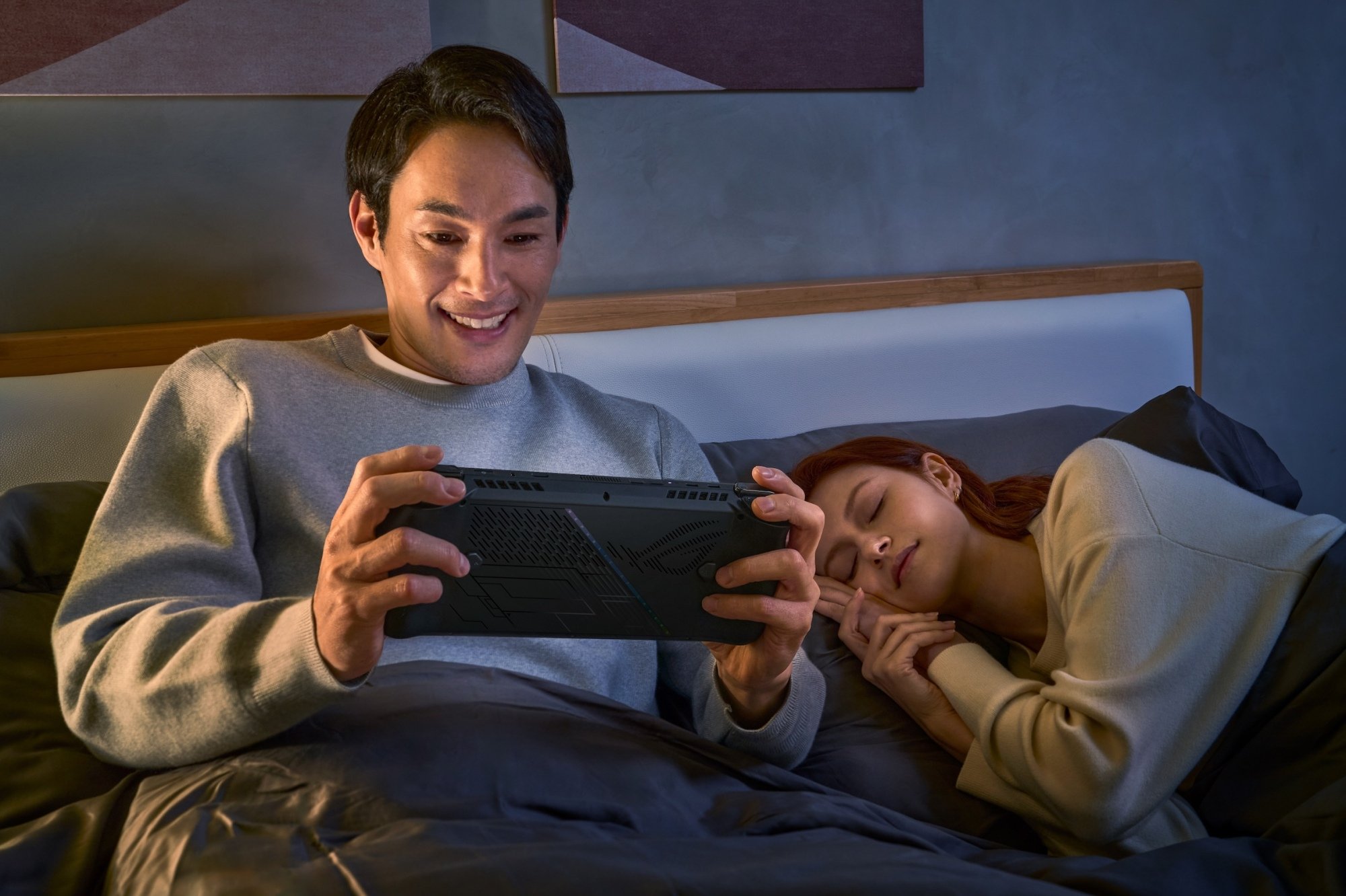 Man in bed holding an Asus ROG Ally X while a woman sleeps next to him